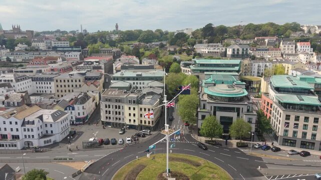 St Peter Port Guernsey .Slow forward drone footage of the weighbridge and roundabout at the foot of St Julian&rsquo;s Avenue over the mast flying flags and flagship office buildings on bright sunny day