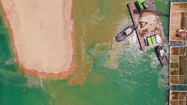 Top-down aerial view of tugboats maneuvering a crane barge during a river dredging operation, with sediment creating a sandbar and contributing to land reclamation along the industrial coastline