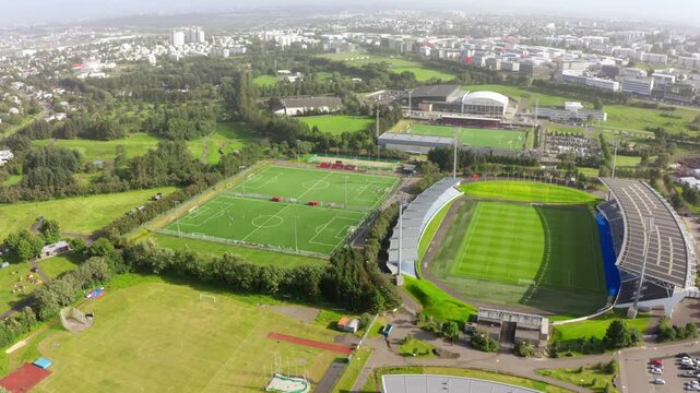 Aerial drone view flying over multiple green soccer fields and a large stadium in a park area with the city of Reykjavik, Iceland in the background.