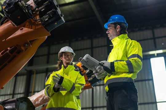 Man and women engineer and worker remote control standing in industrial factory near large manufacturing robotic arm wearing high visibility uniform checking advanced technology during production.