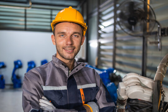 Confident male engineer wearing yellow hard hat standing near blue robot arm equipment designed for automated factory production.