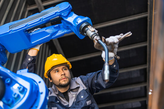 man wearing yellow helmet controls blue robot arm for heavy industrial manufacturing inside modern factory setting showing intense concentration on his daily mechanical work