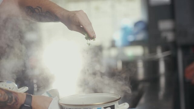 Chef sprinkles fresh herbs into a steaming pot in restaurant kitchen