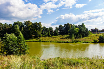 Fototapeta premium Serene countryside pond reflecting fluffy clouds under a bright blue sky, framed by lush green trees and wild grasses in a peaceful summer landscape, idyllic rural nature scenery
