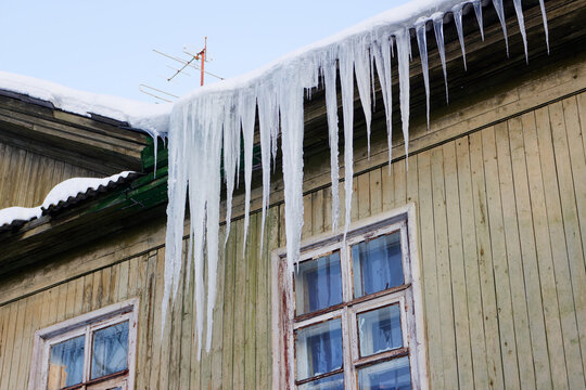 Sharp dangerous icicles hang from the snowy roof edge of an old rustic wooden house during a freezing winter day, creating a beautiful yet menacing frozen natural architecture display