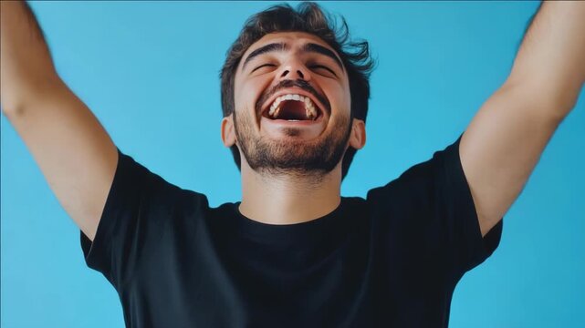 Young man laughs joyfully against blue background. Eyes closed, arms raised high in celebration. Beard, dark hair, black t-shirt, vibrant backdrop. Emotionally expressive, joyful, energetic moment