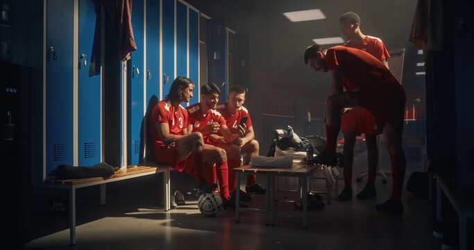 Five Multiethnic Male Soccer Players Watching Smartphone Together in Locker Room: Three Athletes Sit on Bench Smiling at Social Media Content While Two Teammates Lean In, Feeling Amused Before Match
