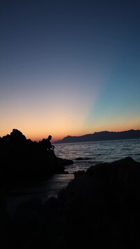 Vertical screen Sunset fishing silhouette on rocky shoreline captures peaceful solitude as an angler waits for the final bite beneath a glowing evening sky by calm sea