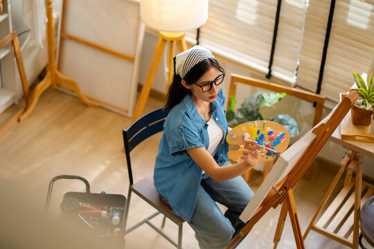 Young Asian woman artist painting on canvas easel with paintbrush in art studio, representing creative professional women designer, fine arts student lifestyle and freelance illustrator
