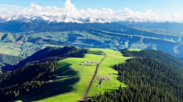 Aerial view of a nomadic yurt camp on rolling green hills with a snow-capped mountain range in the background, Kalajun Grassland, Xinjiang, China.
