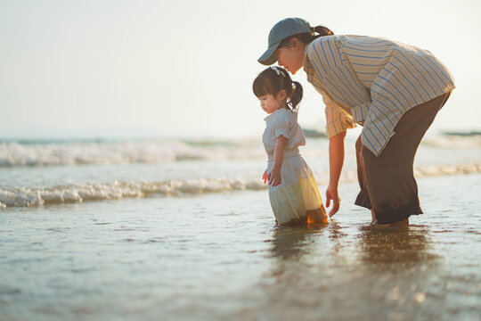 Happy Asian mother and cute little daughter playing in sea water on sandy beach at sunset, family enjoying tropical ocean travel vacation during golden hour, child development, parenting and summer