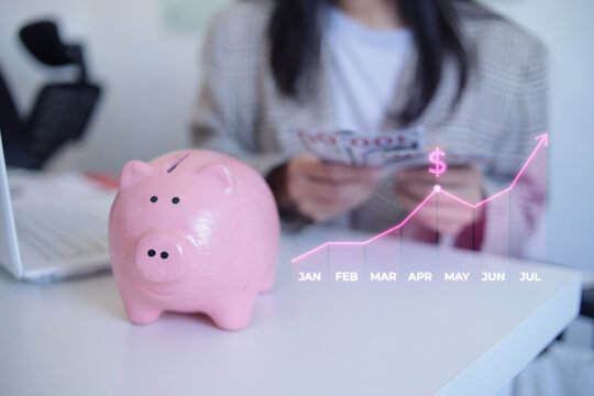 Female Investor Handling Cash Next To Piggy Bank And Rising Financial Hologram On Desk
