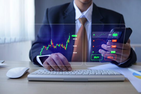 Businessman Using Phone And Keyboard To Track Investment Hologram At Workspace