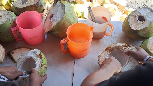 Hands scooping young coconut flesh and pouring coconut water into colorful cups. Top view of street vendor preparing fresh tropical drinks on table.