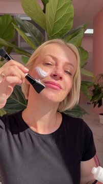 Mature woman applying face cream, facial serum by brush during her daily morning Skincare Routine on cheek at home. Healthy face skin care beauty, anti age concept, skincare application, UGC video