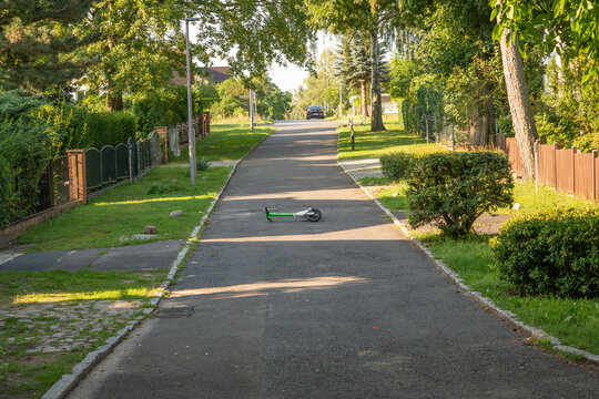 A green electric scooter rests on a shaded path lined with tall trees and garden fences. A parked car and distant houses complete this quiet residential scene.