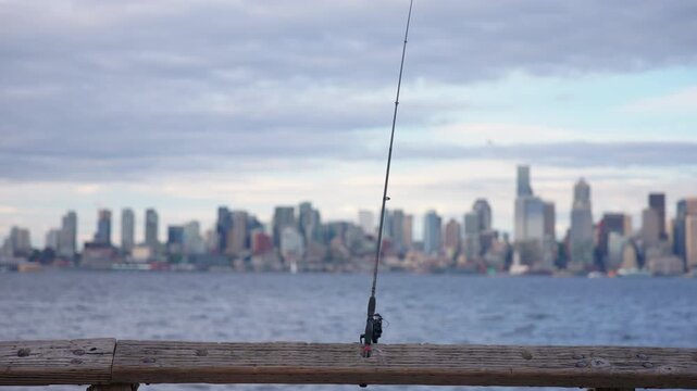 A fishing rod on a pier railing stays in sharp focus while the Seattle skyline blurs across Elliott Bay, with a slow, subtle tilt up revealing the rod's tip, Tilt Up