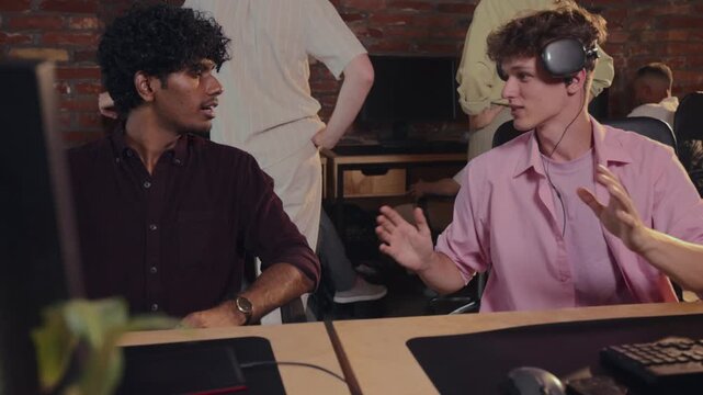 Two young male coworkers discussing work at computers as manager joins conversation. Concept of peer collaboration, workplace communication, team supervision.