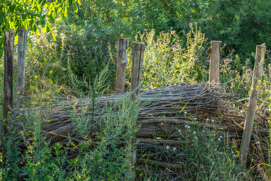 A woven brushwood habitat pile rests between rustic wooden posts surrounded by blooming wildflowers. This dead hedge structure shelters insects and small wildlife in a natural garden.