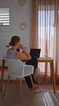 A person sits at a small desk in a home office working on a laptop. Sunlight comes through the curtains. A glass of green drink is beside them.