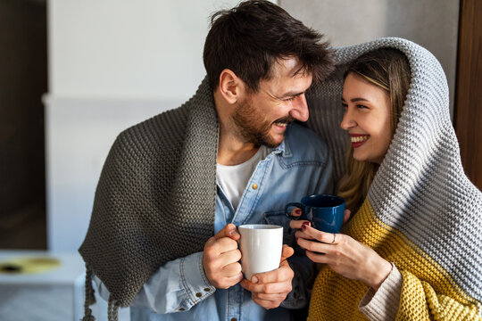 Cheerful young couple covered in blanket embracing while holding cups of tea at home