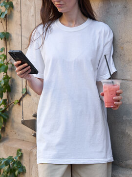 Woman in white t-shirt holding a pink drink and smartphone outdoors