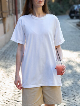 Woman in oversized white t-shirt and shorts holding a pink drink outdoors