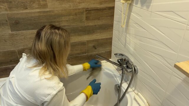 Woman in rubber gloves wiping white bathtub corner with rag in bathroom