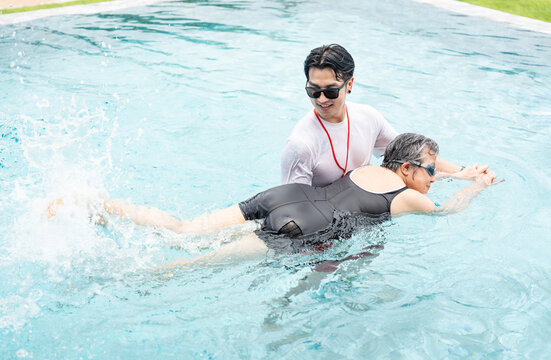 A young asian man instructing a senior woman swimming in the pool, elderly aquatic health exercise concept