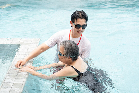 A young asian man instructing a senior woman swimming in the pool, elderly aquatic health exercise concept