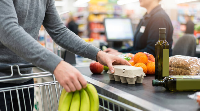 A man places bananas and an egg carton on the checkout counter. In the background, a cashier assists another customer with various items. The store appears to be busy