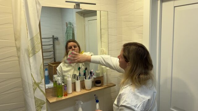Housewife doing domestic chores cleaning glass surface with a towel focused on her reflection in the bathroom mirror interior