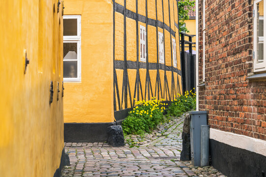 Layered building heritage lines a narrow cobbled lane between yellow half timbered facade and red brick wall in Ribe for Danish architecture, local history and travel themes.