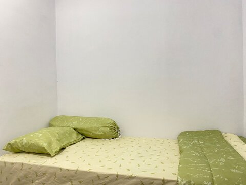 Simple cozy bedroom corner with a neatly made bed, green pillows, and light yellow sheets against a plain white wall