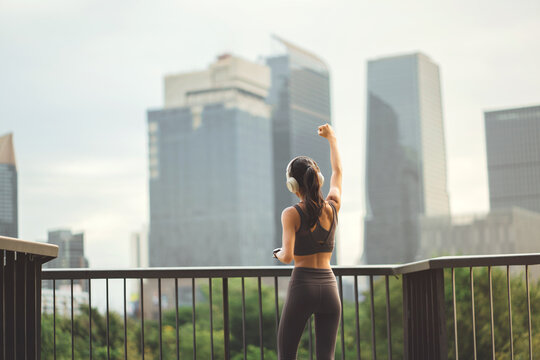 Back view Asian sportswoman raising arm in fist pump celebrating success while standing at city park bridge, fit female runner wearing headphones for urban running workout motivation.