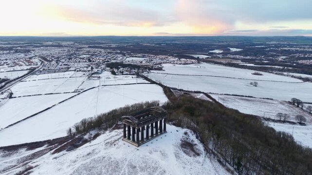 Elevated wide-angle drone orbit shot around the snow-covered Penshaw Monument in Sunderland, revealing a vast panorama of County Durham under a winter sunset sky