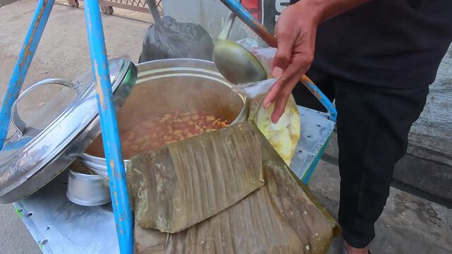Video of Street vendor serving soup from large pot. Hand stirs stew with wooden spoon. Food items over banana leaves. Vendors wear dark clothing and sandals. 4K Footage of everyday street food scene.