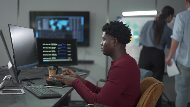 Young African male software engineer writing program code on multiple desktop screens while his colleagues working in the background in a modern office.