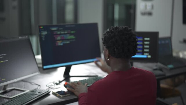 Over-the-shoulder shot of young African male software engineer writing program code on multiple desktop screens while his colleagues working in the background in a modern office.
