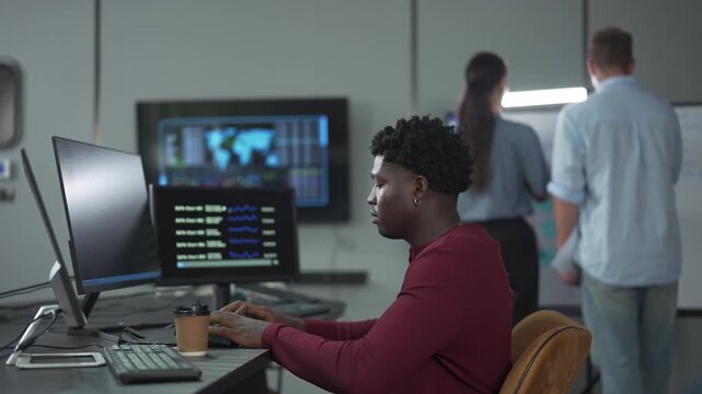 Young African male software engineer writing program code on multiple desktop screens while his colleagues working in the background in a modern office.
