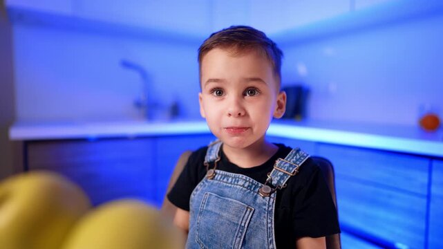 Close up portrait of boy behind yellow apples. He looks toward the camera as cool kitchen lights blur across the background.