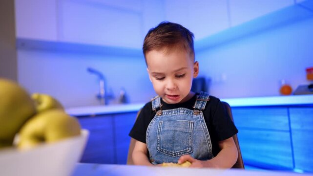 Boy holding bitten apple at kitchen table. He looks down at the fruit while a white bowl sits blurred in the foreground.