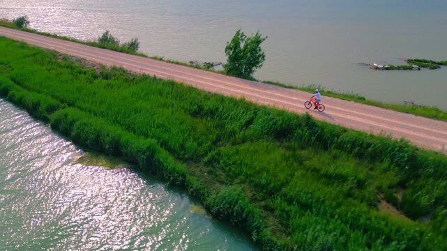 A senior adult enjoys a peaceful cycling journey along a scenic river, embracing an active lifestyle. The aerial shot captures their movement against lush greenery and calm waters.