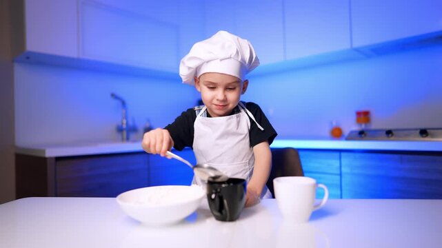 Young boy chef serving food with ladle. He wears a white hat and apron beside a bowl and two cups on the table.