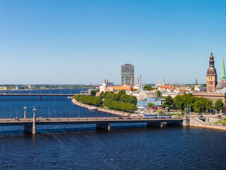 Obraz premium Aerial view of the historic waterfront of Riga Old Town along the Daugava river, with the embankment, tree-lined promenade and the towers of the medieval centre, under a clear blue summer sky.