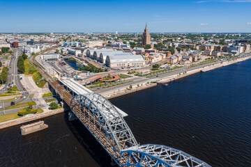 Aerial drone view of the five vaulted pavilions of Riga Central Market, built from former zeppelin hangars, beside the railway bridge and the Daugava river, under a clear blue summer sky. © Aerial Film Studio