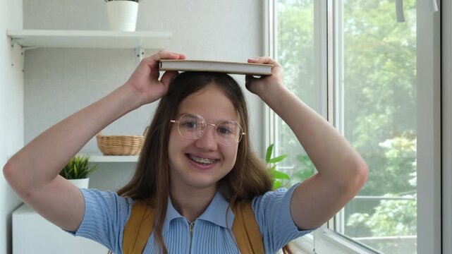 Smiling teen student girl with backpack holding book on head. Happy female teenager in glasses with dental braces looking at camera