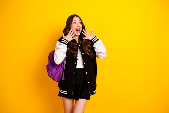 Excited female student with backpack and varsity jacket expressing surprise against bright yellow background