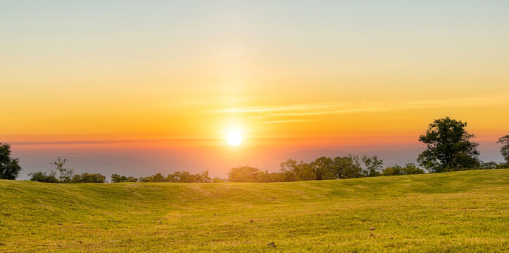 Golden sunset glowing over a green grass field with silhouetted trees and layered mountains fading into the horizon.