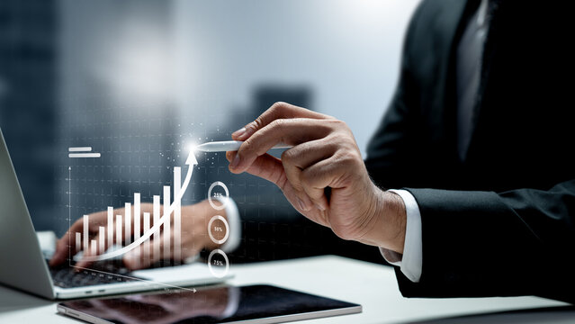 Businessman pointing a stylus at a glowing digital growth chart rising from a laptop, symbolizing financial success. Concept of business growth or finance